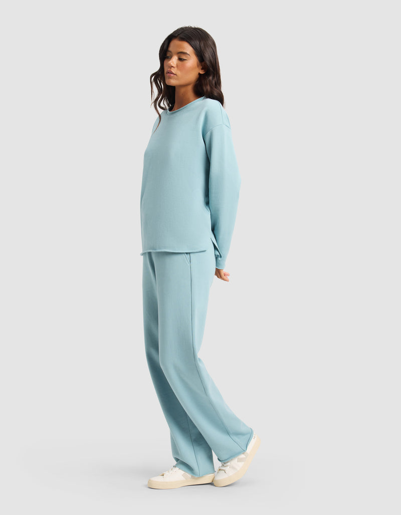 A woman models the Cozy Earth Women's Chelsea Set—loose-fitting, light blue long-sleeve top and matching wide-leg pants—paired with white sneakers, standing sideways against a plain light background.