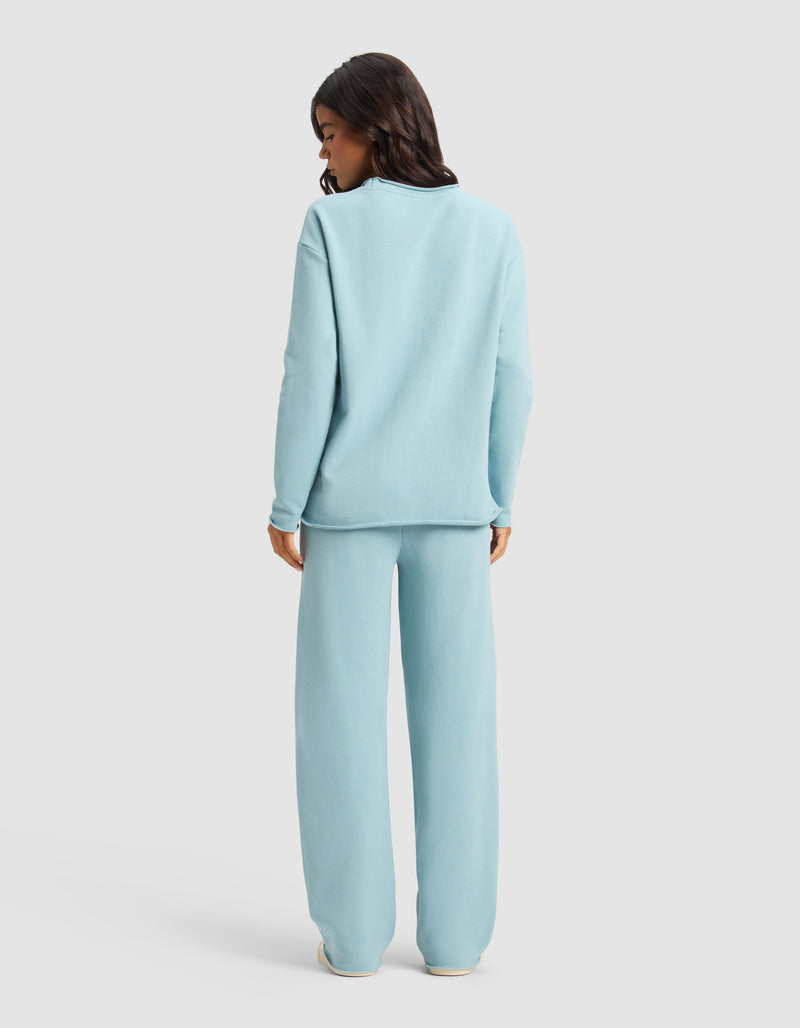 A woman faces away, wearing the Cozy Earth Women's Chelsea Set—loose-fitting light blue sweatshirt and matching wide-leg pants—against a plain light background.