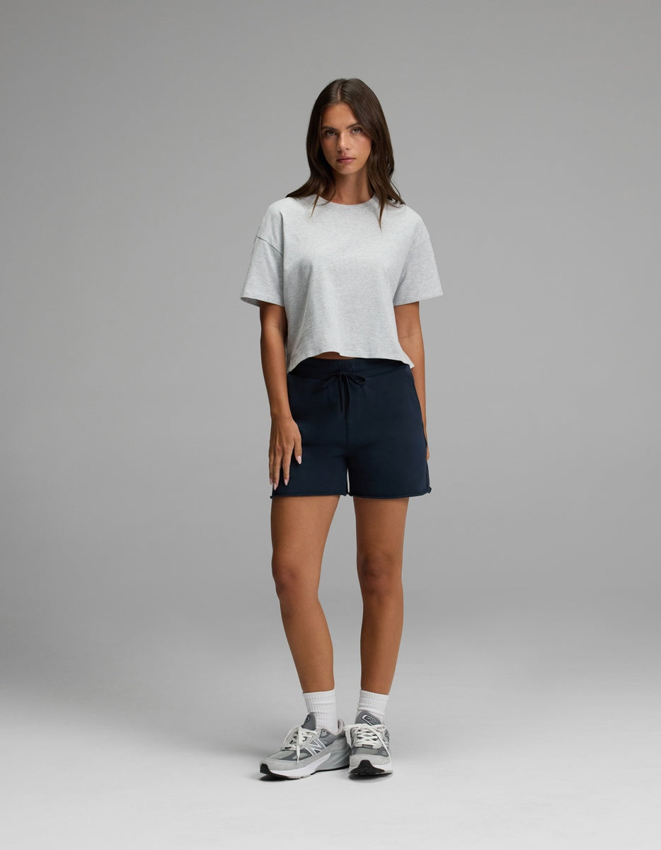 A woman stands against a plain background, wearing Cozy Earth’s Women’s Chelsea Short, paired with a light gray T-shirt and gray sneakers. She has long brown hair and a neutral expression, with her hands relaxed by her sides.