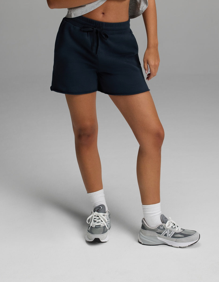 A person wearing the Cozy Earth Women’s Chelsea Short in Eclipse, a gray cropped top, white socks, and gray New Balance sneakers stands on a light gray surface. Their face is not visible in the image.|Color:Eclipse