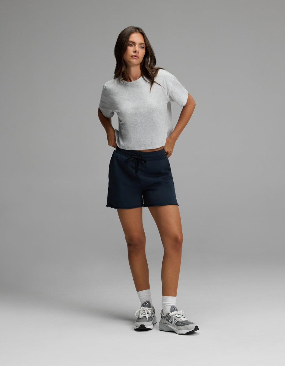 A woman stands against a plain backdrop wearing Cozy Earth’s Women’s Chelsea Short paired with a light gray short-sleeve t-shirt, white socks, and gray sneakers. Her hands rest on her hips as she gazes slightly to the side.