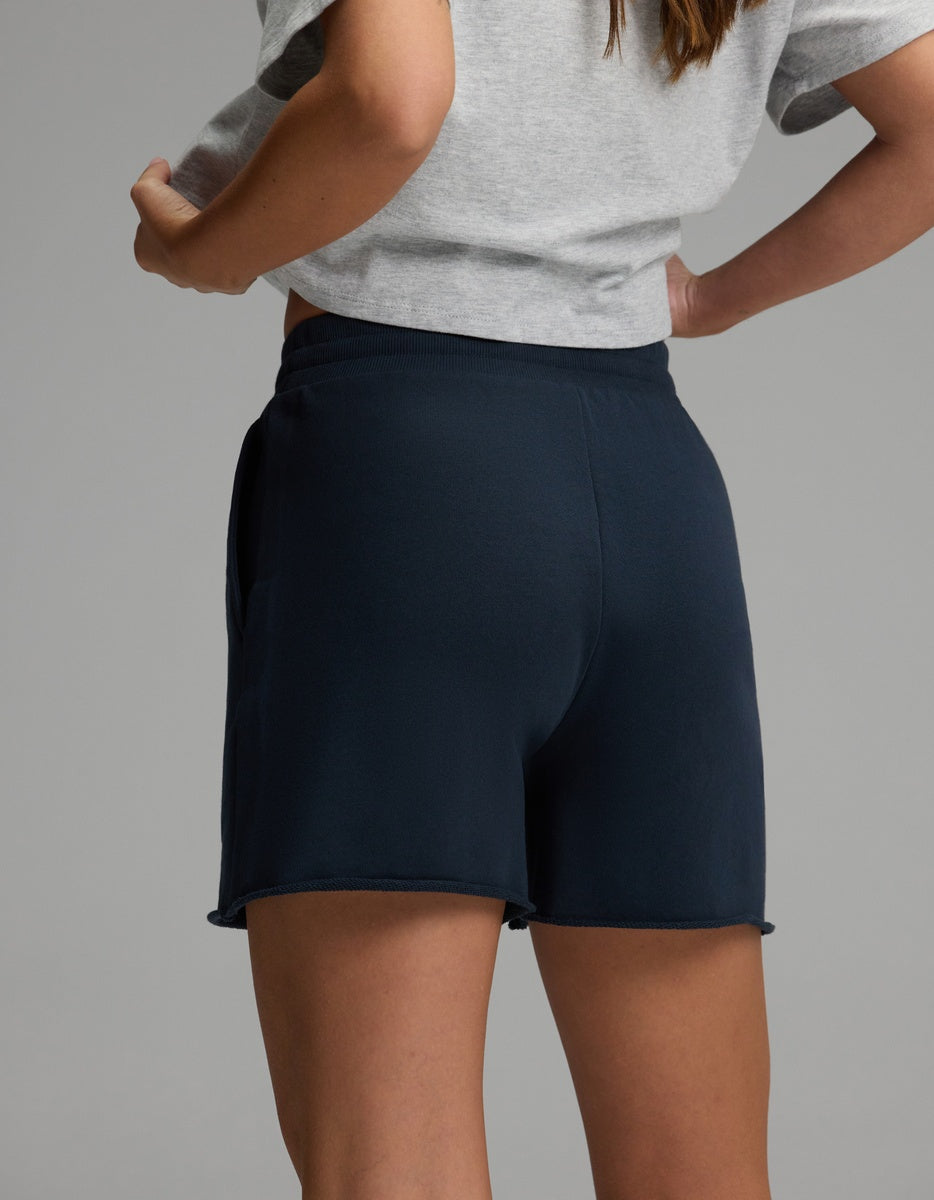 A person is shown from the back and waist down, wearing Cozy Earth’s Women’s Chelsea Short in dark blue with a gray t-shirt, standing against a plain gray background.