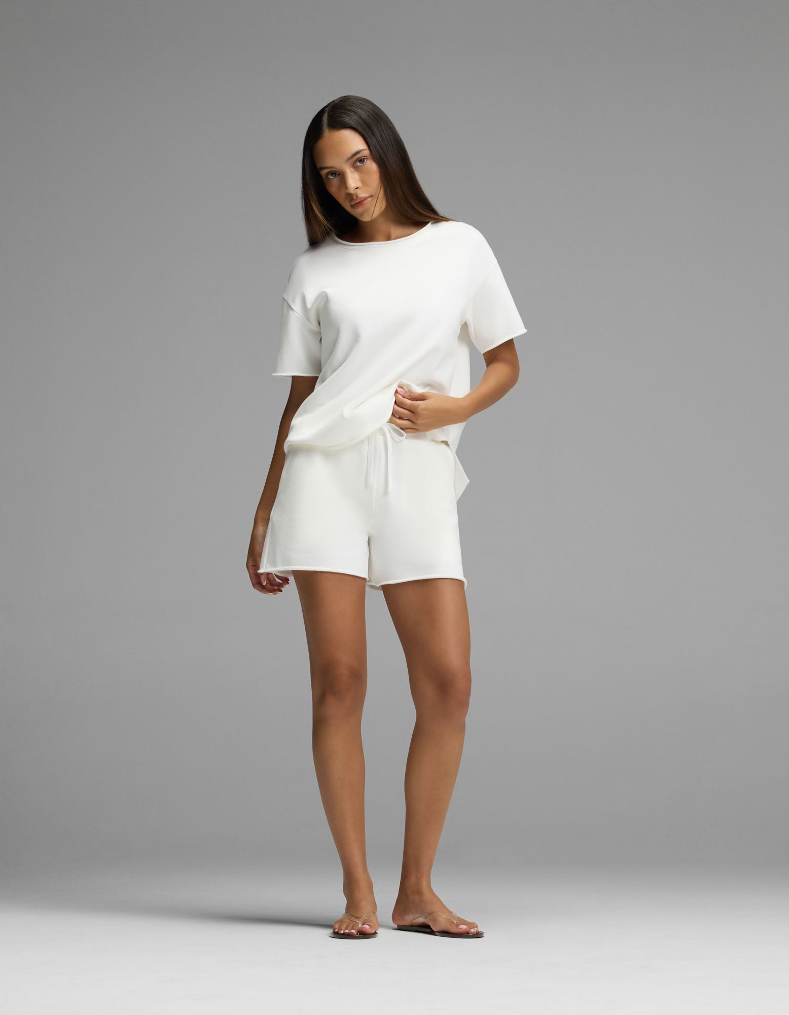 A woman stands barefoot against a gray background, wearing Cozy Earth’s Women’s Chelsea Short Sleeve Set in white, with her right hand resting in her pocket.
