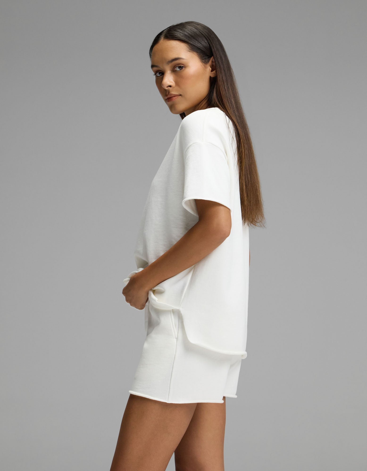 A woman with long straight hair stands sideways against a plain gray background, hands in the pockets of her Cozy Earth Women’s Chelsea Short Sleeve Set—a loose white T-shirt paired with matching white shorts.|Color:Bone