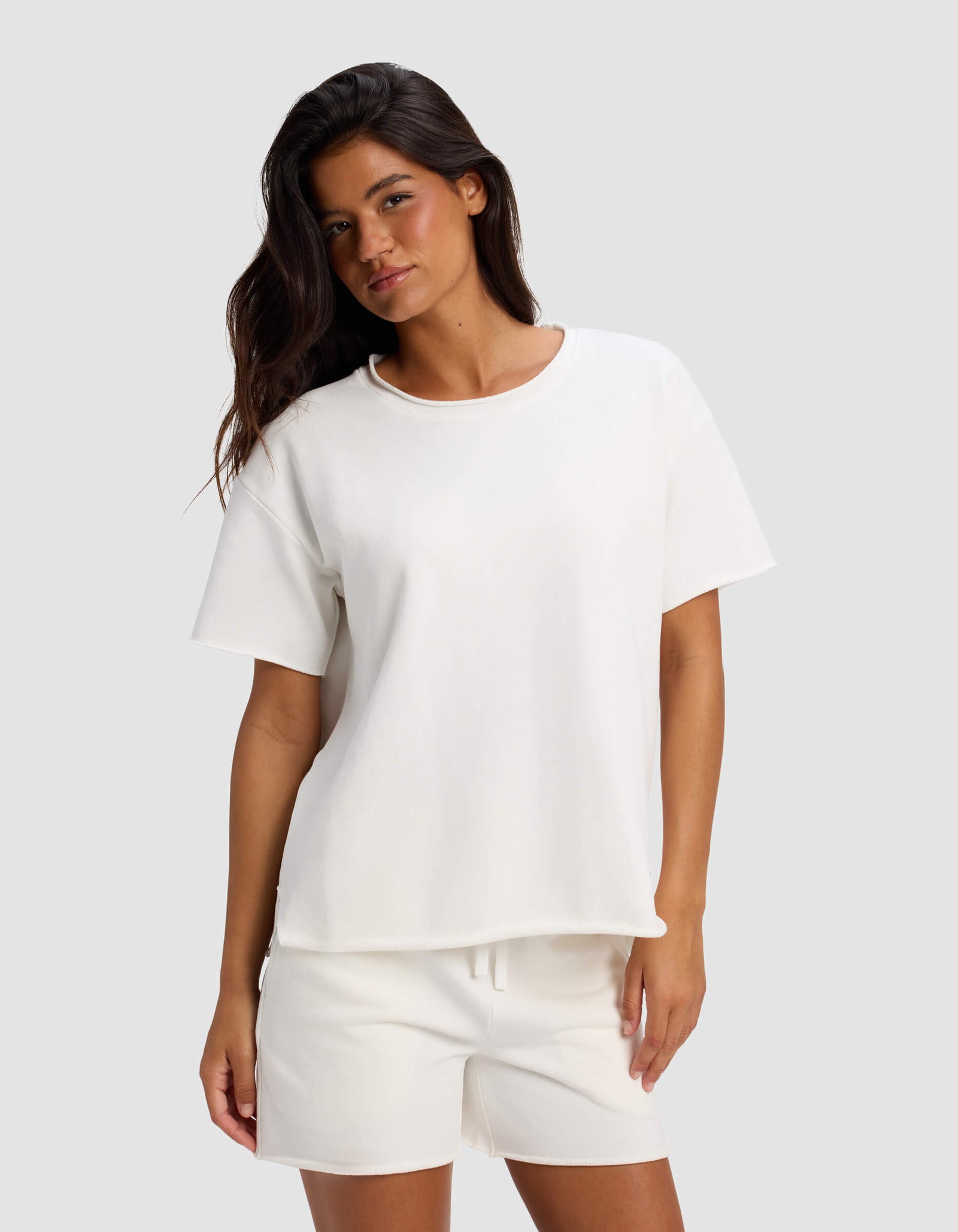 A woman with long dark hair models the Women's Chelsea Short Sleeve by Cozy Earth, paired with white shorts. She stands against a light gray background in a relaxed, casual pose with a slight smile.