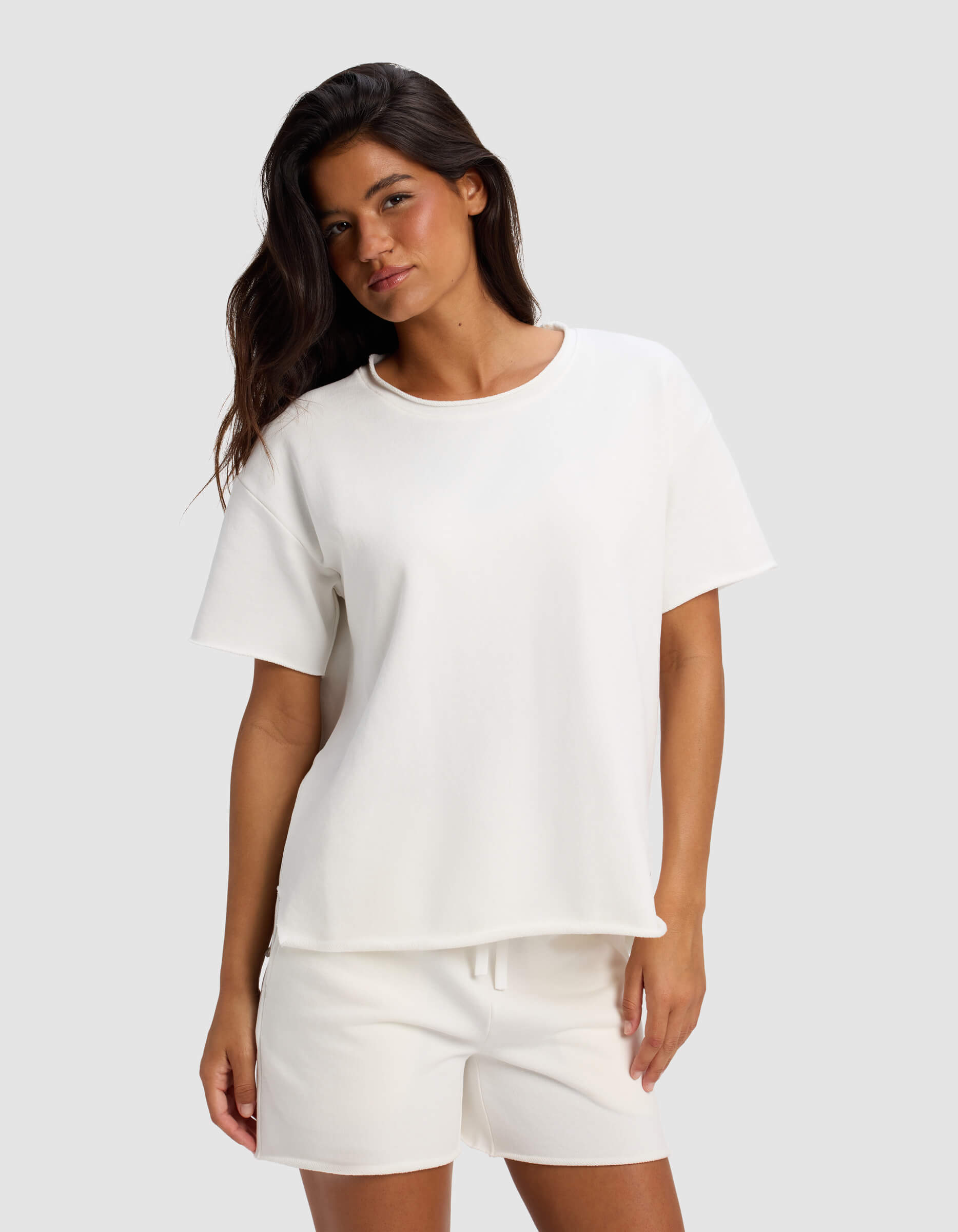 A woman with long dark hair models the Women's Chelsea Short Sleeve by Cozy Earth, paired with white shorts. She stands against a light gray background in a relaxed, casual pose with a slight smile.