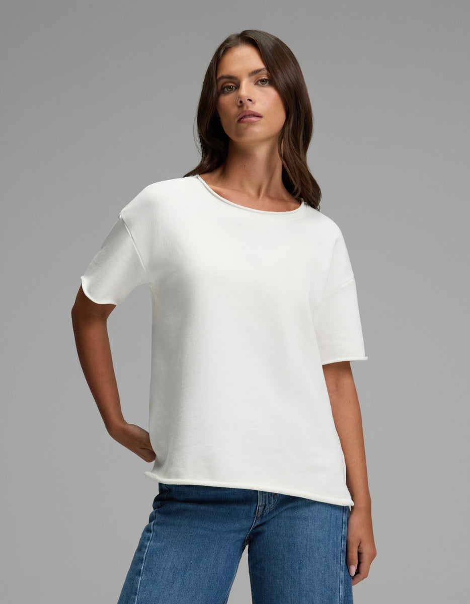 A woman with long brown hair stands against a neutral background, wearing the Cozy Earth Women’s Chelsea Short Sleeve in Bone and blue jeans. She has one hand in her pocket and a confident expression.|Color:Bone