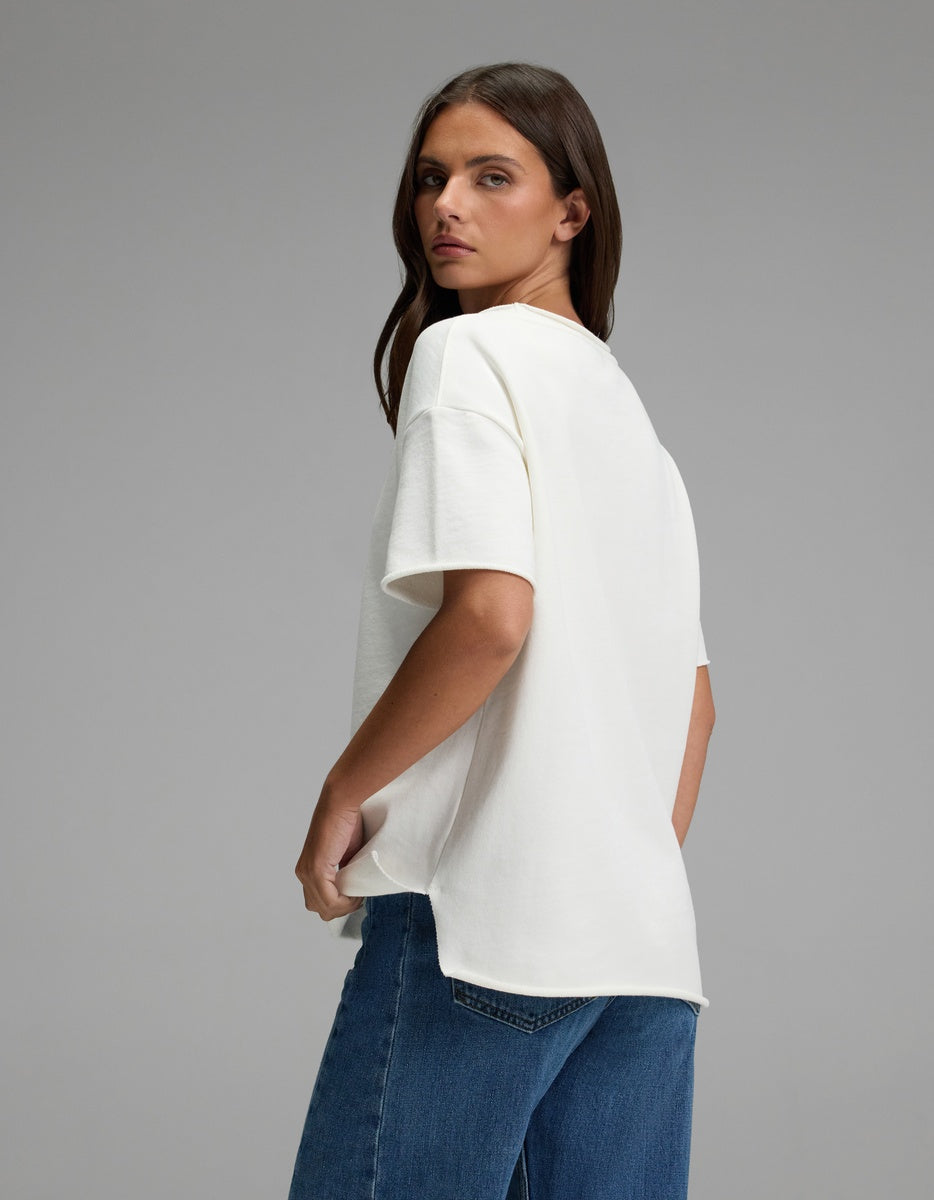A woman with long brown hair, dressed in the Cozy Earth Women’s Chelsea Short Sleeve and blue jeans, stands against a plain gray background, glancing over her shoulder at the camera.|Color:Bone