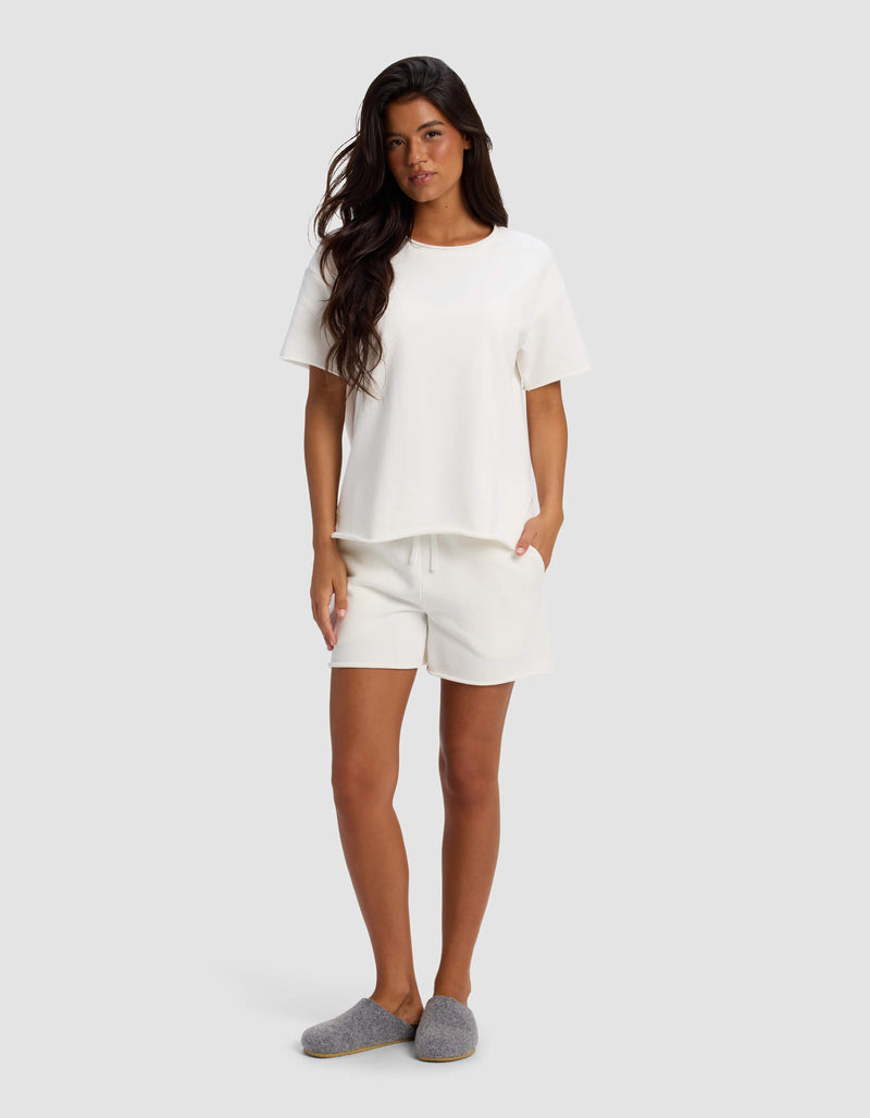 A woman stands against a plain light gray background wearing Cozy Earth’s Women's Chelsea Short Sleeve top and matching white shorts, with her hands in her pockets and gray slippers on her feet.