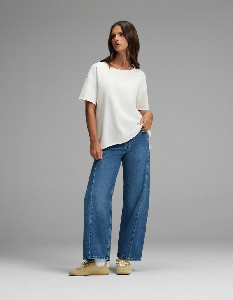 A woman stands against a gray background wearing the Cozy Earth Women’s Chelsea Short Sleeve, wide-leg blue jeans, and beige sneakers, with one hand in her pocket and looking to the side.
