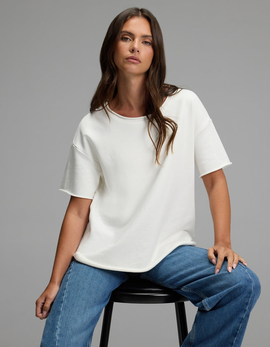 A woman with long brown hair, wearing the Cozy Earth Women’s Chelsea Short Sleeve and blue jeans, sits on a black stool against a plain gray background, looking confidently at the camera.