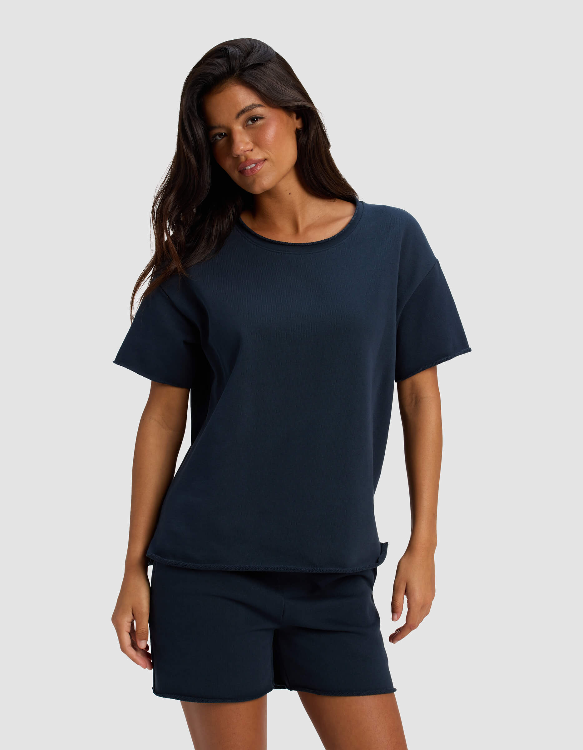 A woman with long dark hair wears Cozy Earth's Women's Chelsea Short Sleeve navy blue t-shirt and matching shorts, standing against a plain light gray background.|Color:Eclipse