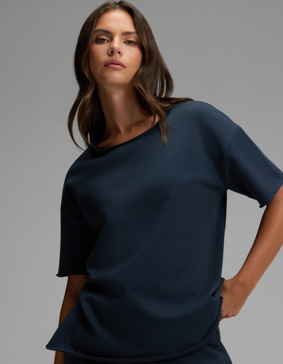 A woman with long brown hair wears the Cozy Earth Women’s Chelsea Short Sleeve in Eclipse, a loose-fitting navy blue top, and poses with one hand in her pocket against a plain gray background.