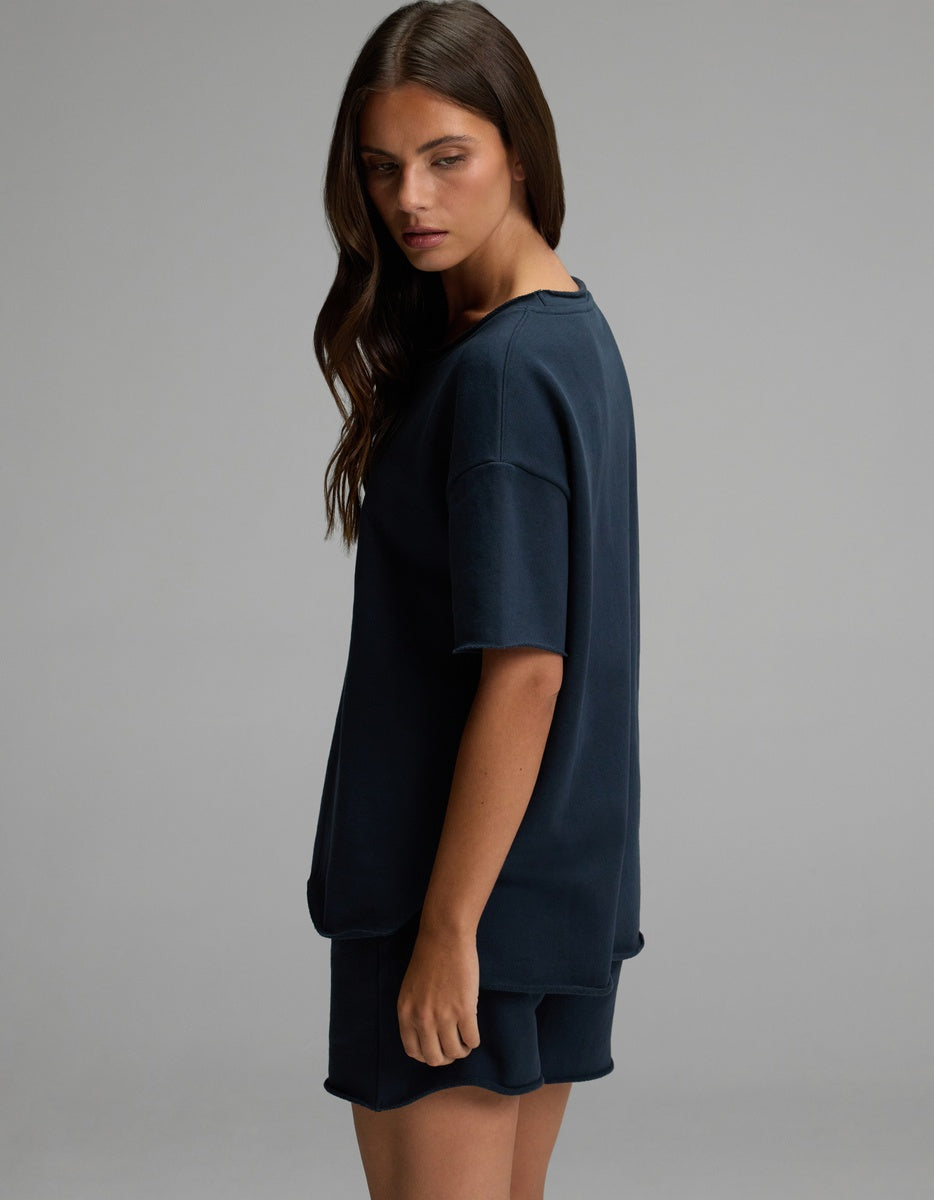 A woman with long brown hair stands sideways against a plain gray background, wearing the Cozy Earth Women’s Chelsea Short Sleeve top and matching shorts in dark blue. 
