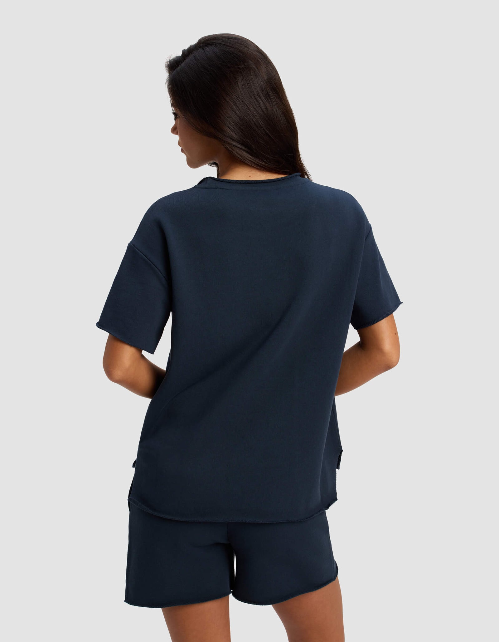 A woman with long dark hair stands with her back to the camera, wearing Cozy Earth’s Women's Chelsea Short Sleeve and matching shorts in navy blue against a plain light gray background.