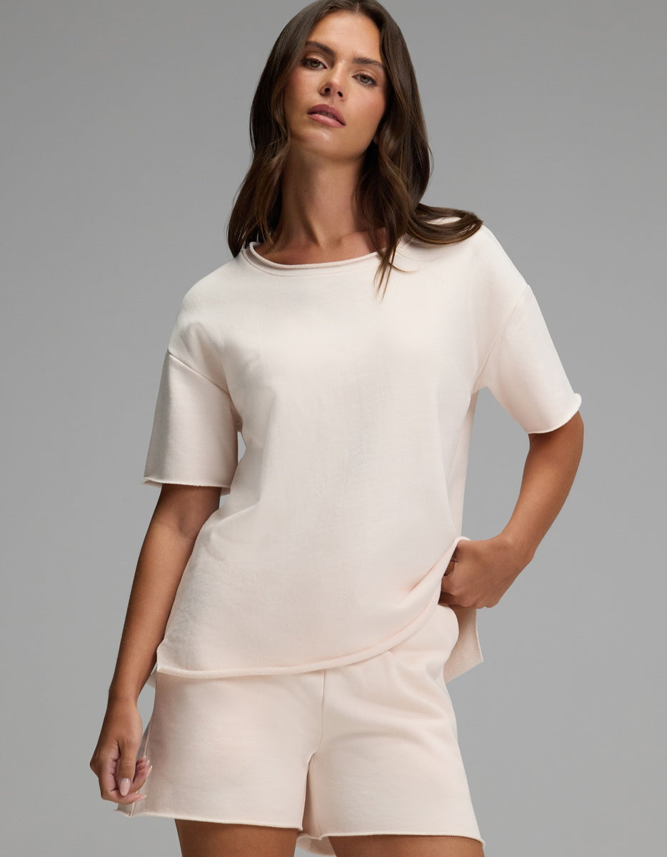 A woman with long brown hair wears the Cozy Earth Women’s Chelsea Short Sleeve in Petal with matching shorts, standing against a plain gray background with one hand in her pocket.