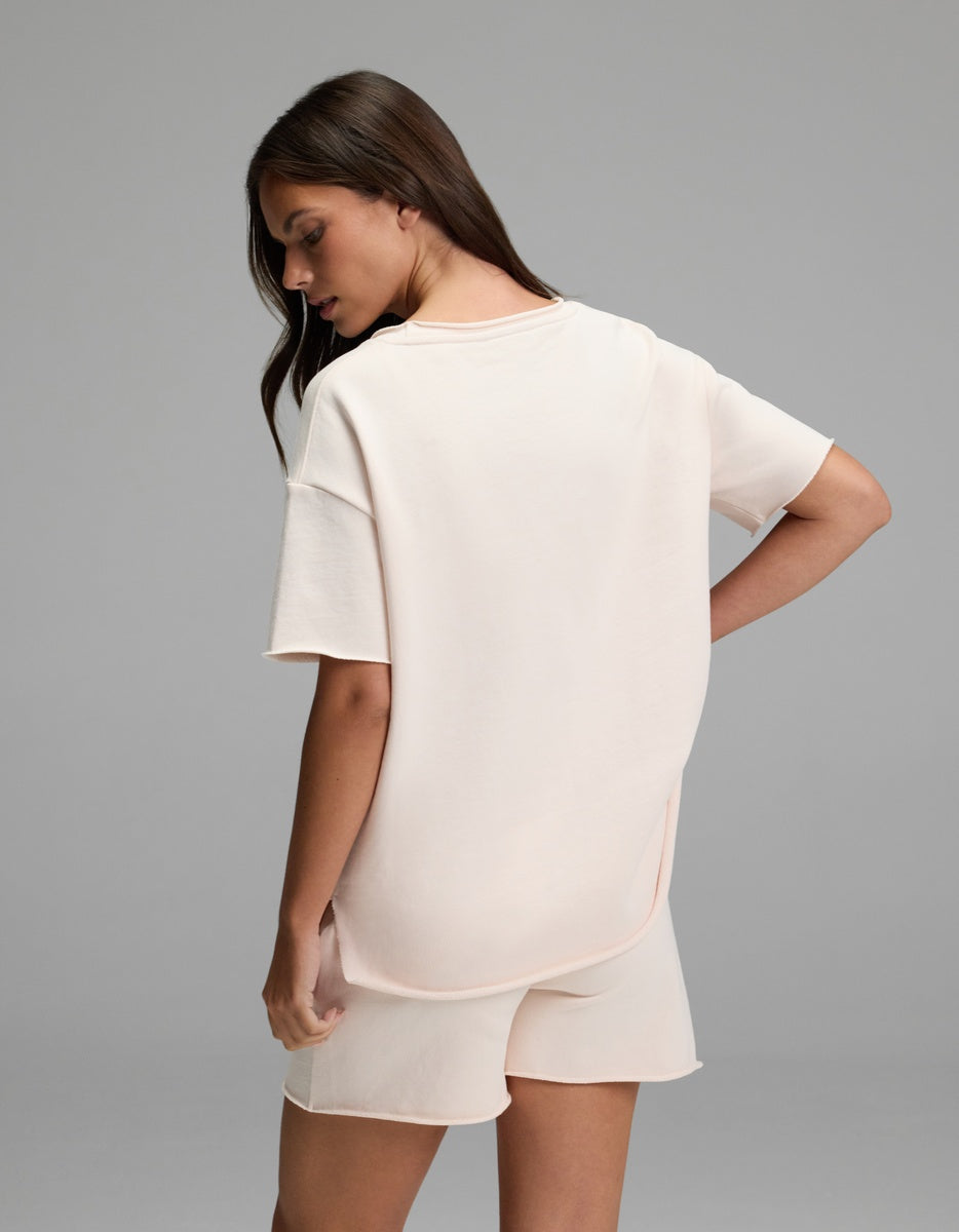 A woman with long brown hair stands sideways, wearing Cozy Earth’s Women’s Chelsea Short Sleeve T-shirt and matching shorts in pale pink, set against a plain gray background.
