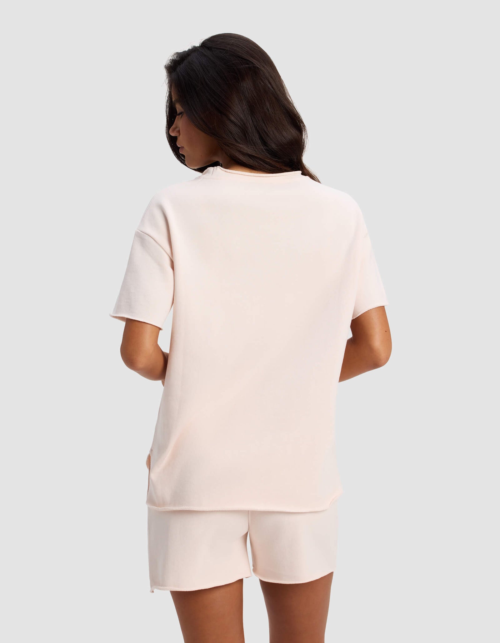 A woman with long dark hair stands facing away, wearing the Cozy Earth Women's Chelsea Short Sleeve shirt and matching shorts against a plain light gray background.