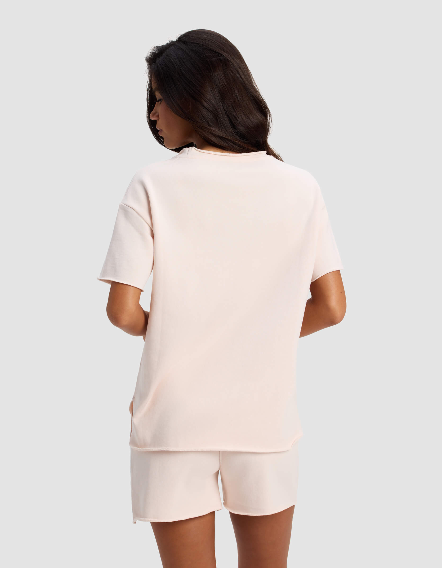 A woman with long dark hair stands facing away, wearing the Cozy Earth Women's Chelsea Short Sleeve shirt and matching shorts against a plain light gray background.