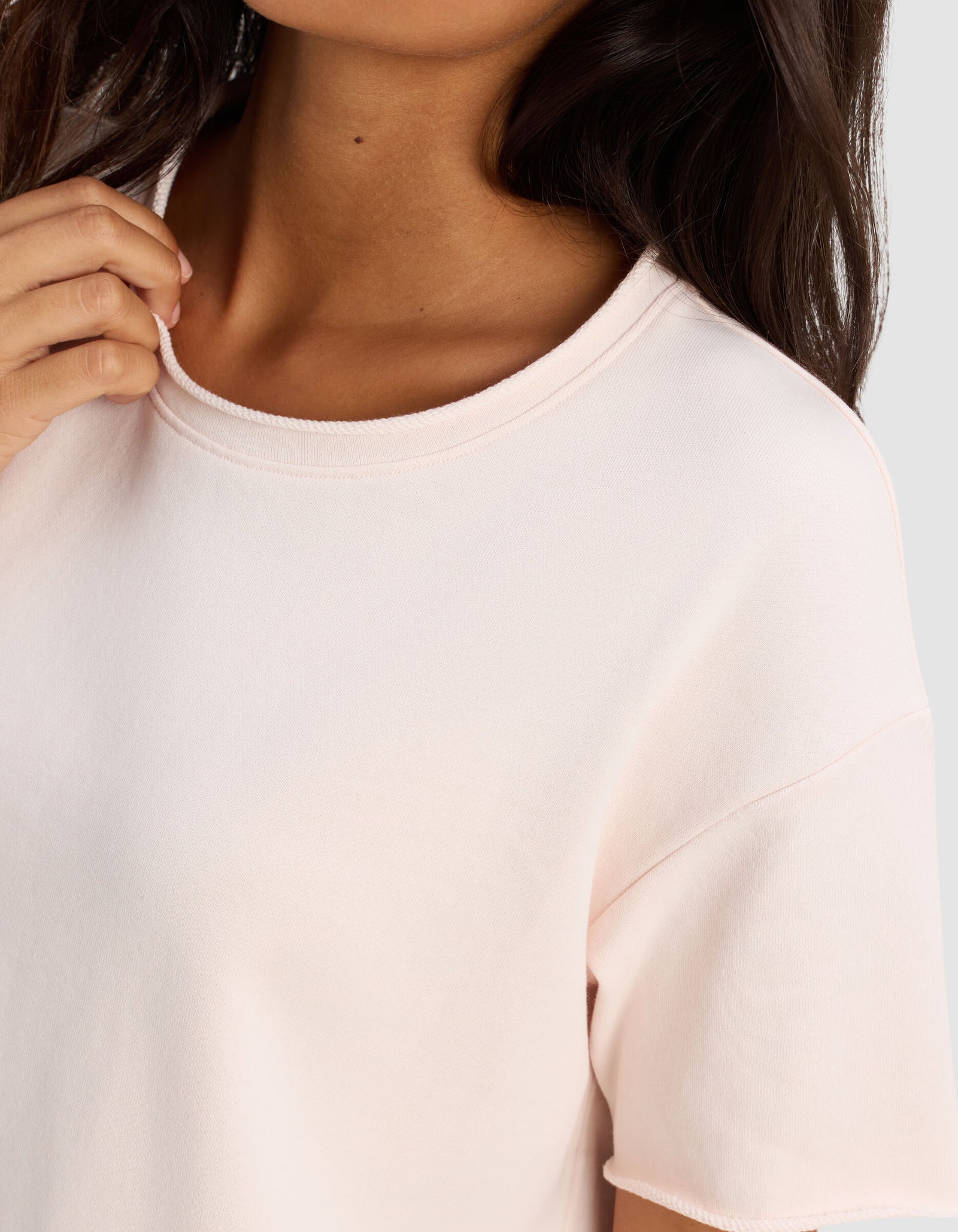 A woman in a Cozy Earth Women's Chelsea Short Sleeve tee in light pink gently holds the neckline. Her dark hair falls over her shoulder, and her face is partially out of frame.