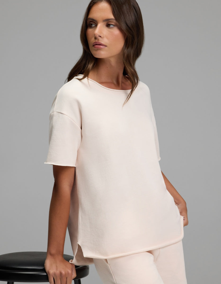 A woman with long brown hair wears the Cozy Earth Women’s Chelsea Short Sleeve top and matching pants in light beige, sitting on a black stool against a plain gray background and gazing to the side.