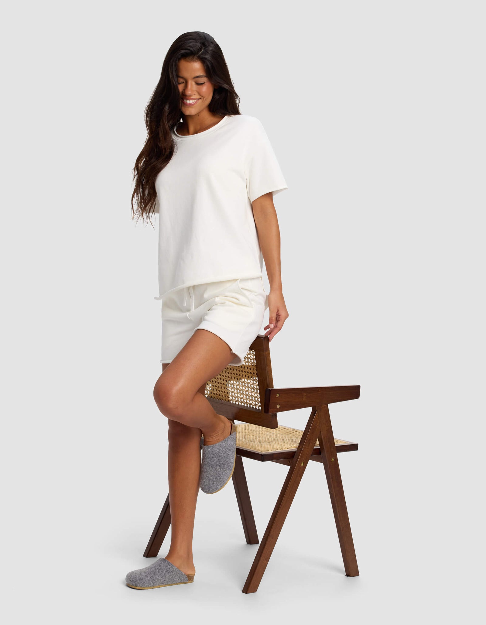 A woman smiles and rests one foot on a wooden chair, wearing Cozy Earth Women's Chelsea Shorts, a white t-shirt, and gray slippers against a plain light gray background.