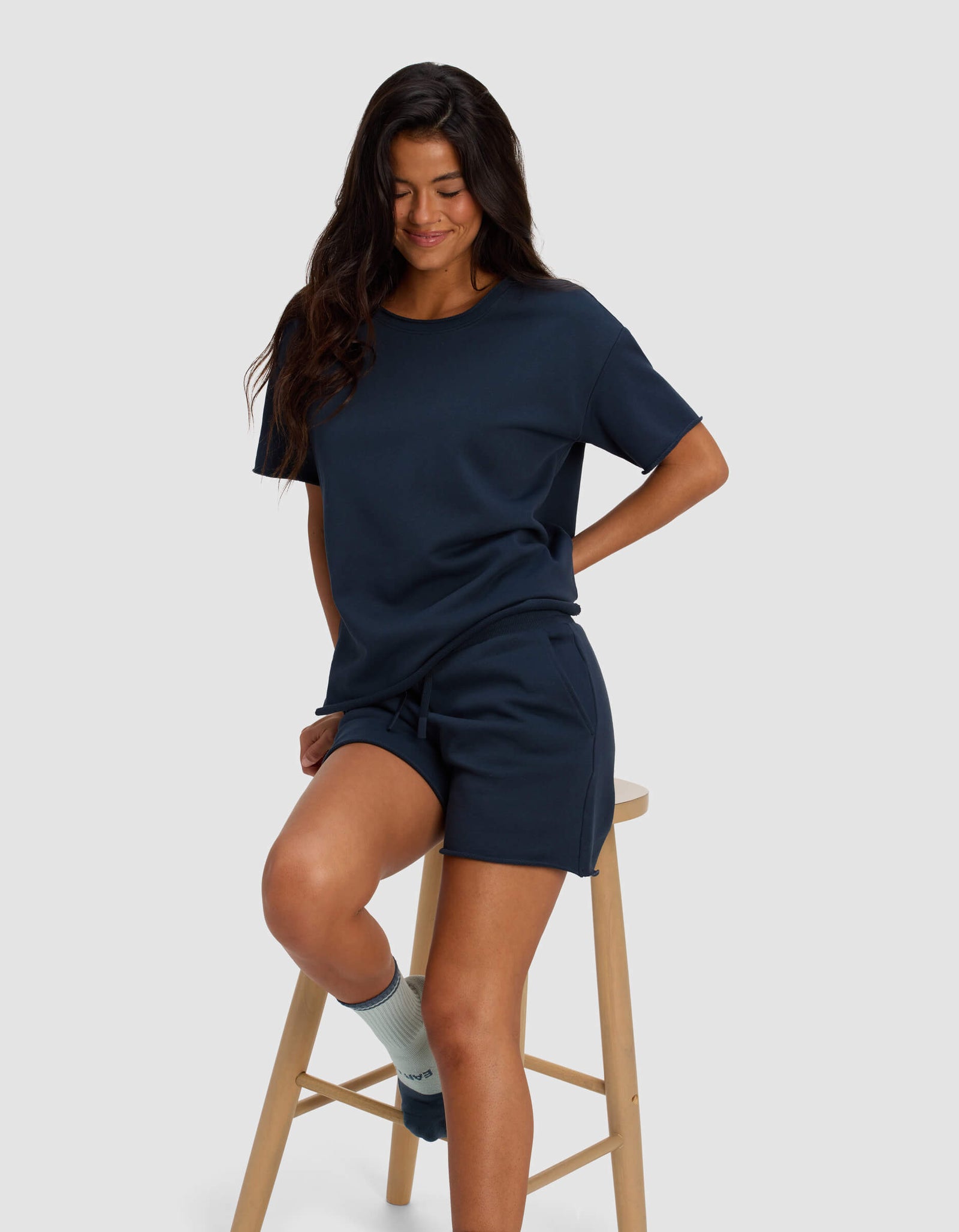 A woman with long dark hair, wearing a navy blue t-shirt and Cozy Earth Women's Chelsea Shorts, sits on a wooden stool against a plain light gray background, smiling with her eyes closed. She is also wearing light blue socks.