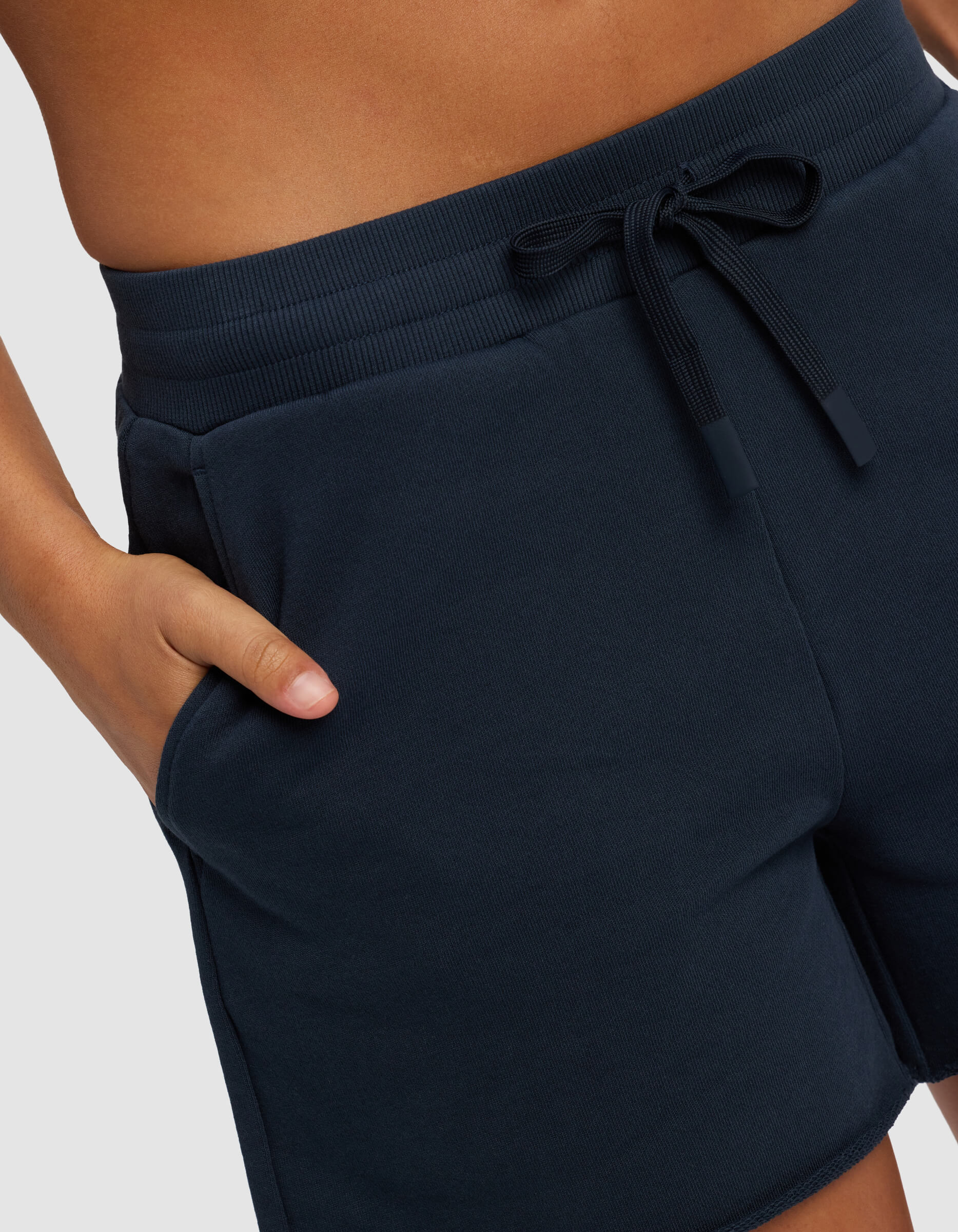 A person wears Cozy Earth's Women's Chelsea Short in dark navy, featuring an elastic waistband and drawstring, standing with one hand in their pocket. The image focuses on the waist and upper thighs against a plain background.