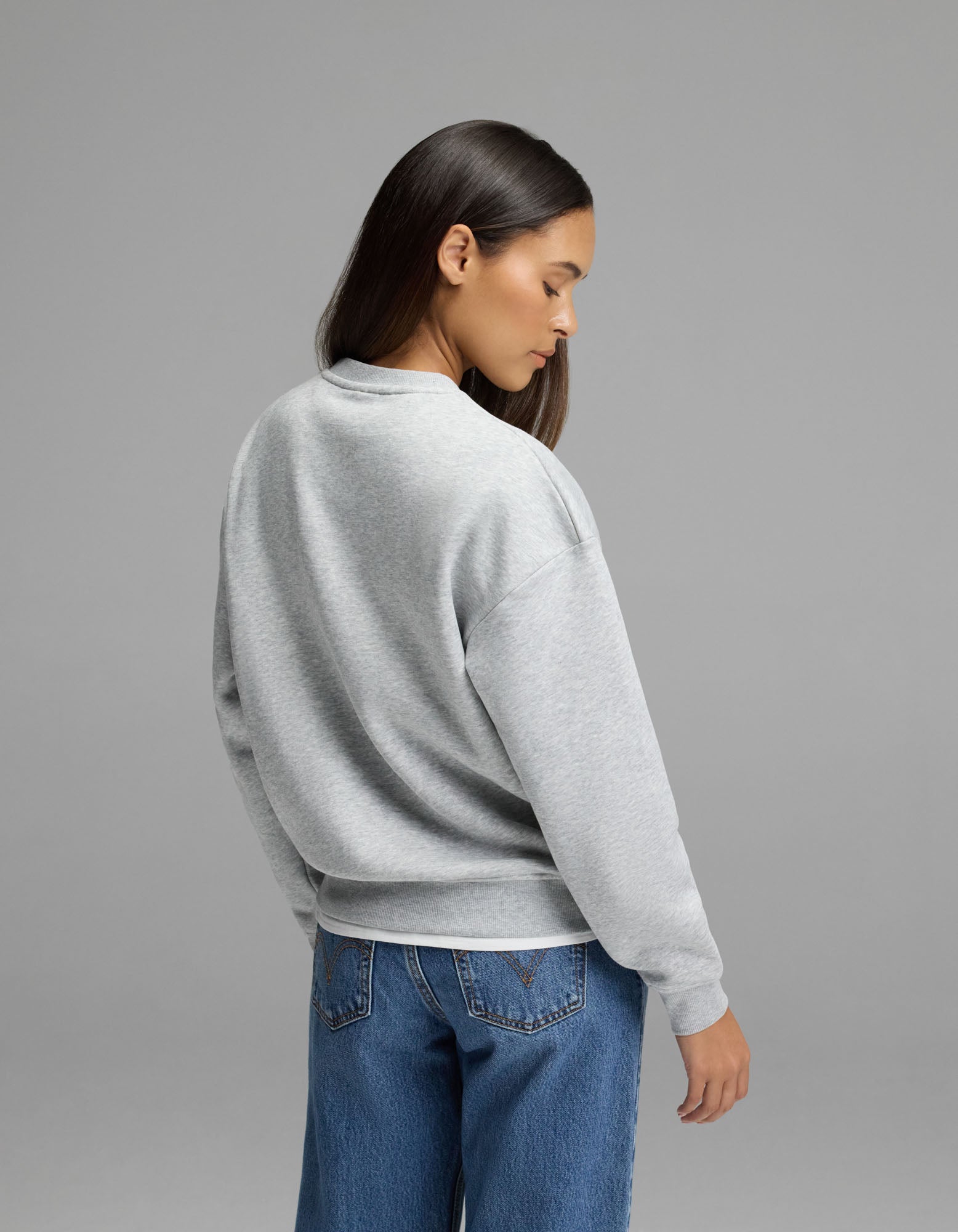 A woman with long dark hair, wearing the Cozy Earth Women’s Cityscape 2.0 Crewneck in light gray and blue jeans, stands facing away from the camera against a plain gray background.|Color:Heather Grey