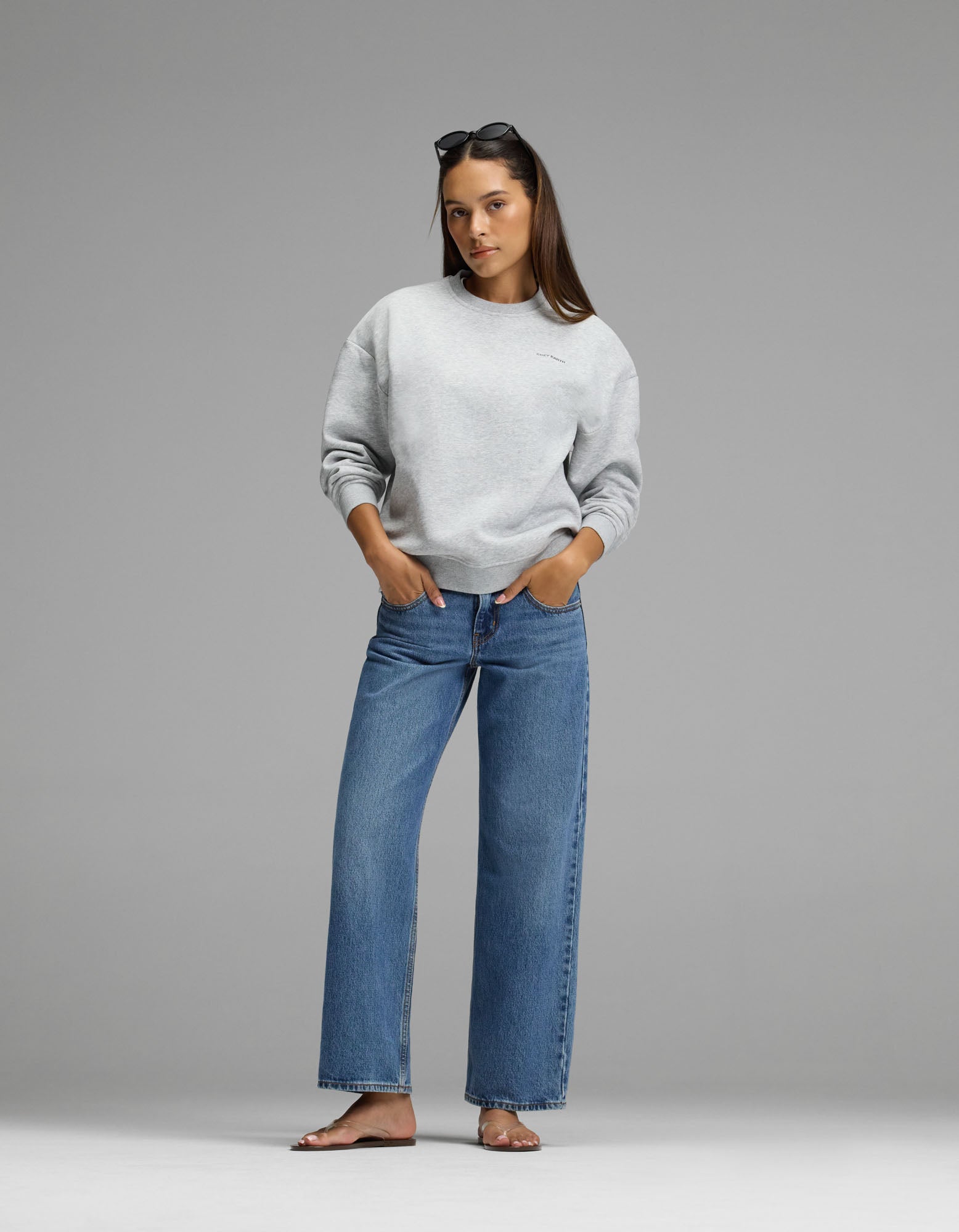 A woman stands against a gray background, wearing the Cozy Earth Women’s Cityscape Crewneck in light gray, paired with blue jeans and sunglasses on her head. Her hands are in her pockets and she is barefoot.