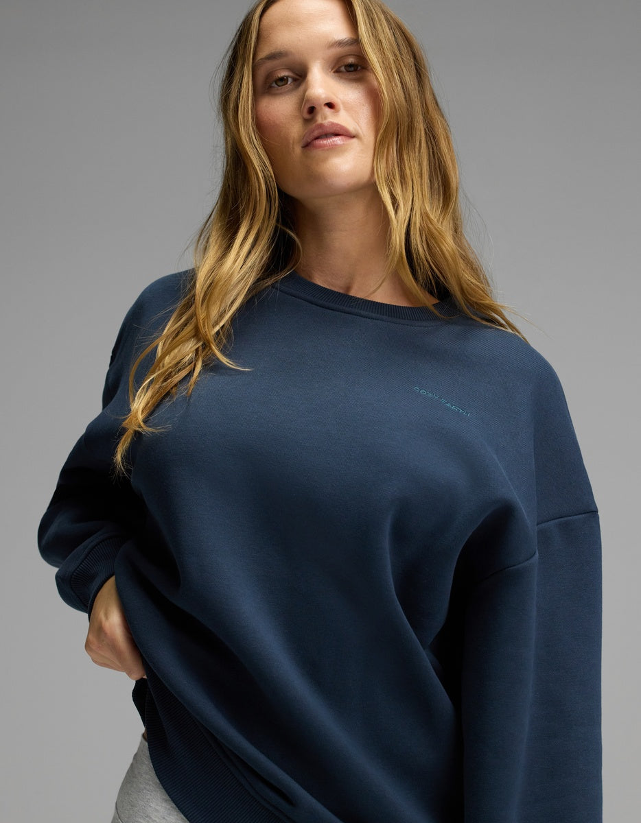 A woman with long, wavy hair stands confidently against a plain gray background, wearing the Cozy Earth Women’s CityScape Crewneck in navy blue paired with light gray pants.