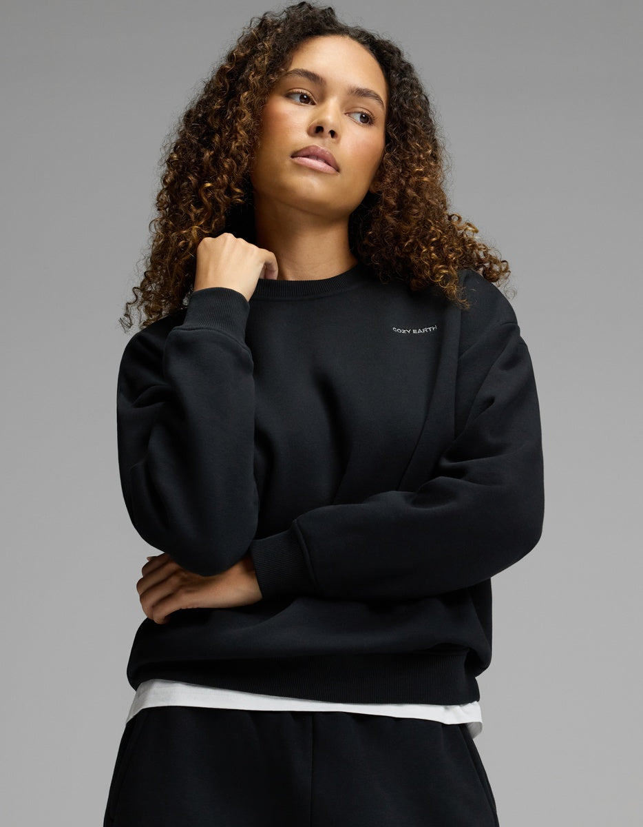 Wearing the Cozy Earth Women’s CityScape Crewneck in black over a white shirt, a woman with curly hair stands against a plain gray background, her right hand touching her chin and her left arm crossed over her chest.