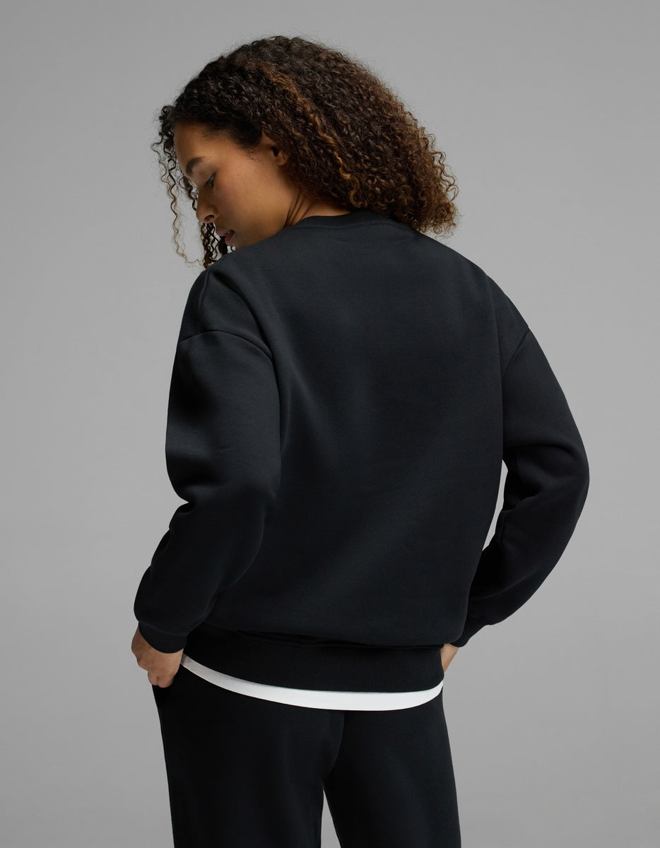 A person with curly hair stands facing away, wearing the Cozy Earth Women’s CityScape Crewneck in black over a white shirt and black pants, set against a plain gray background.