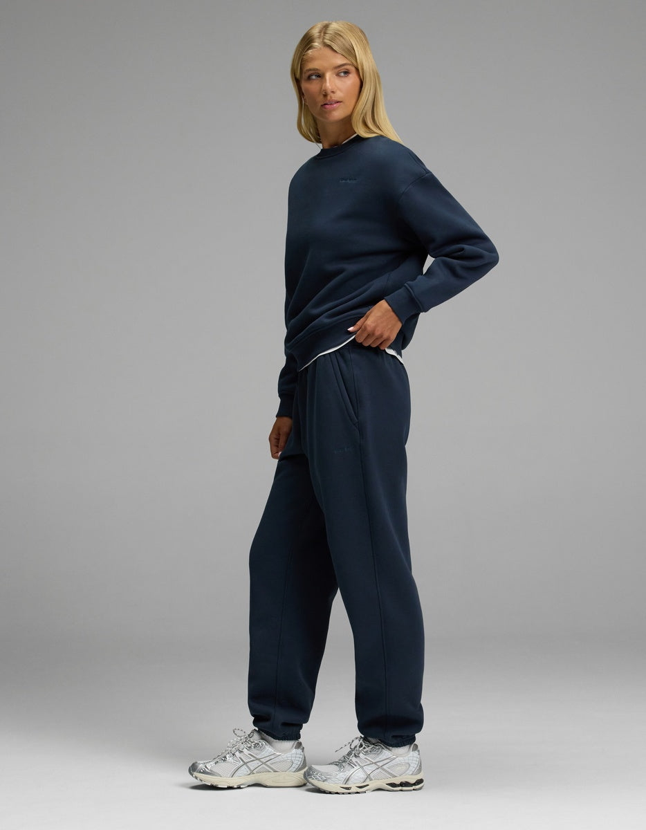 A woman with long blonde hair stands in a relaxed pose against a plain gray background, wearing Cozy Earth’s Women’s CityScape Crewneck & Sweatpant Set in navy blue, paired with white sneakers.