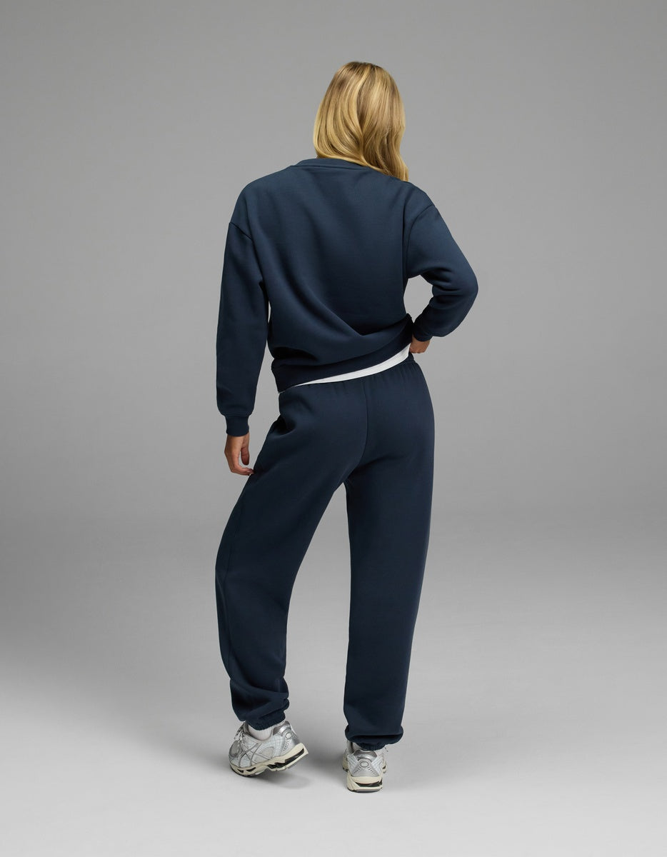 A woman with long blonde hair stands facing away from the camera against a plain gray background, wearing the Cozy Earth Women’s CityScape Crewneck & Sweatpant Set in dark blue, paired with white sneakers.