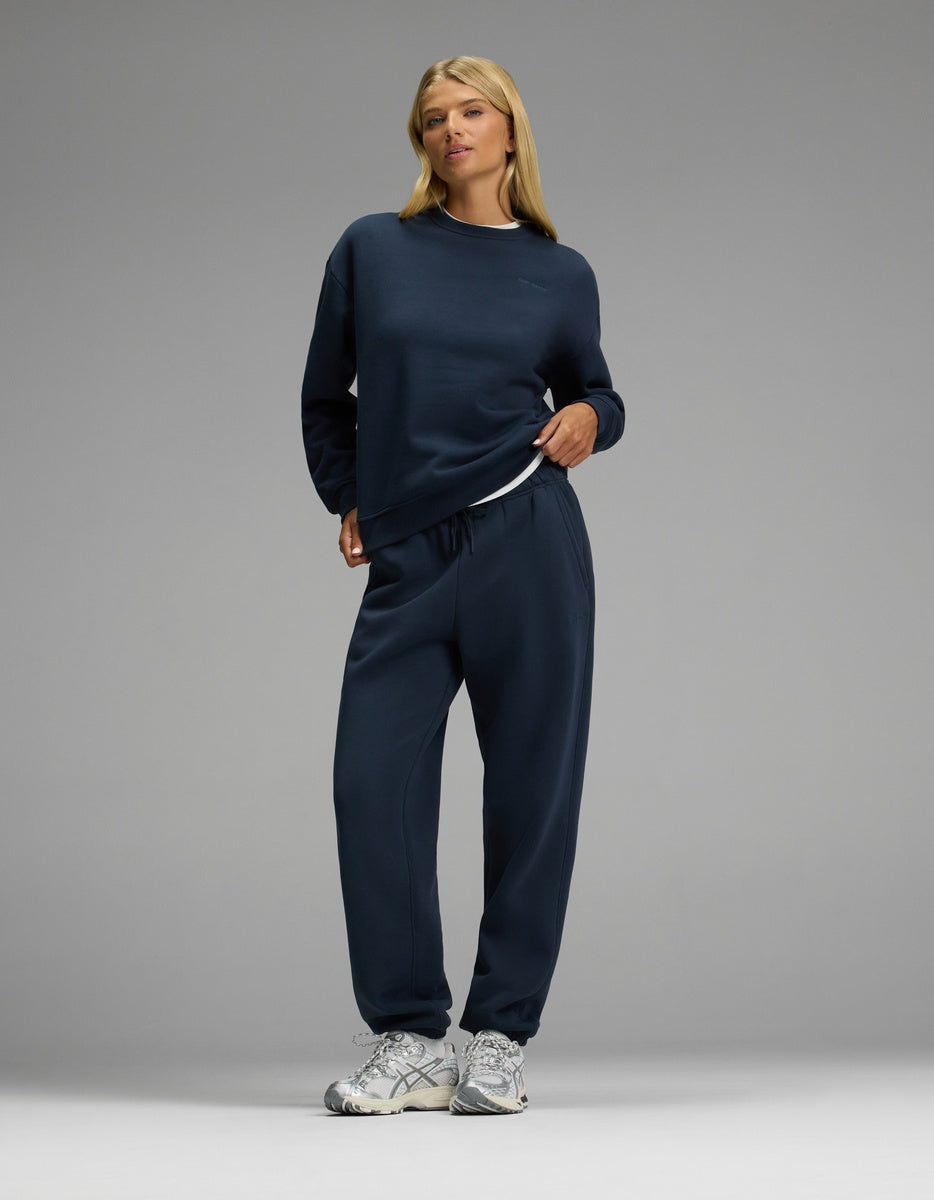 A woman with blonde hair stands confidently against a plain gray background, wearing the Cozy Earth Women’s CityScape Crewneck & Sweatpant Set in navy blue, paired with white sneakers.|Color:Eclipse
