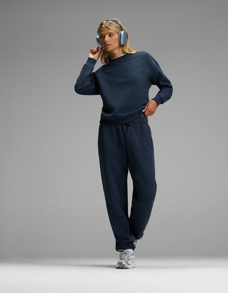 A woman with blonde hair stands against a plain gray background wearing the Cozy Earth Women’s CityScape Crewneck & Sweatpant Set in blue, paired with blue headphones and white and gray athletic shoes.