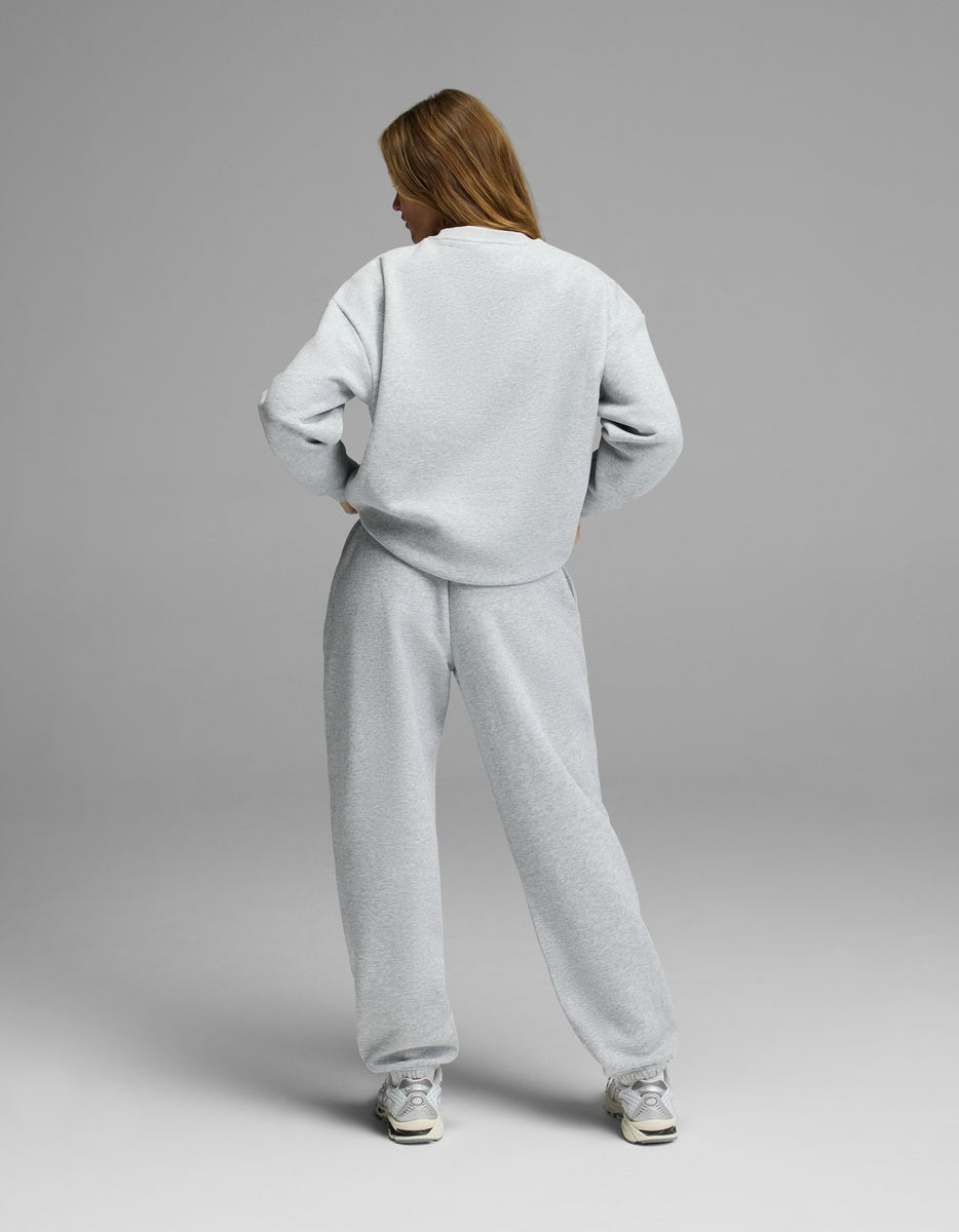 A person stands facing away, wearing the Cozy Earth Women’s CityScape Crewneck & Sweatpant Set in light gray. They have long hair, white sneakers, and their hands rest on their hips against a plain gray background.