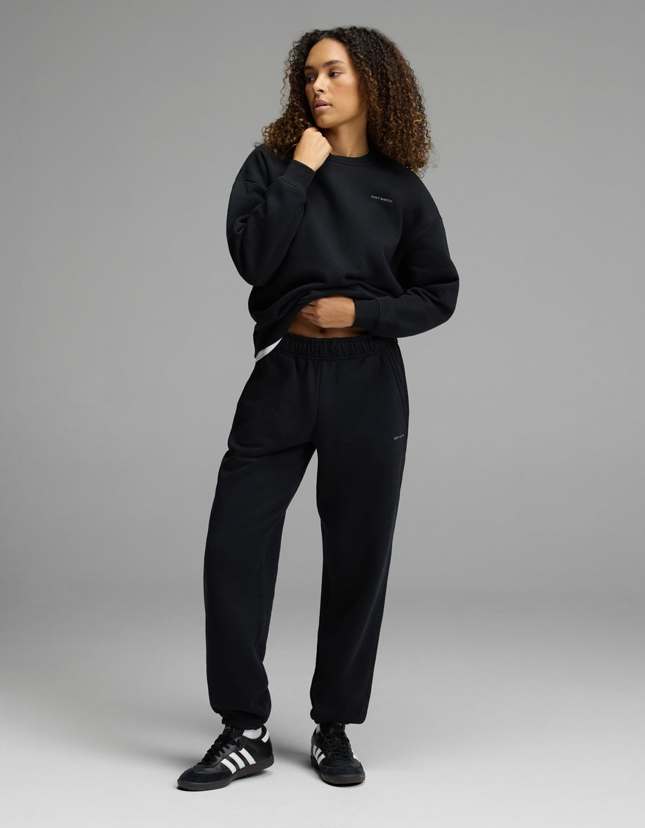 A person with curly hair wears the Women’s CityScape Crewneck & Sweatpant Set by Cozy Earth, standing in a relaxed pose against a plain gray background.|Color:Jet Black