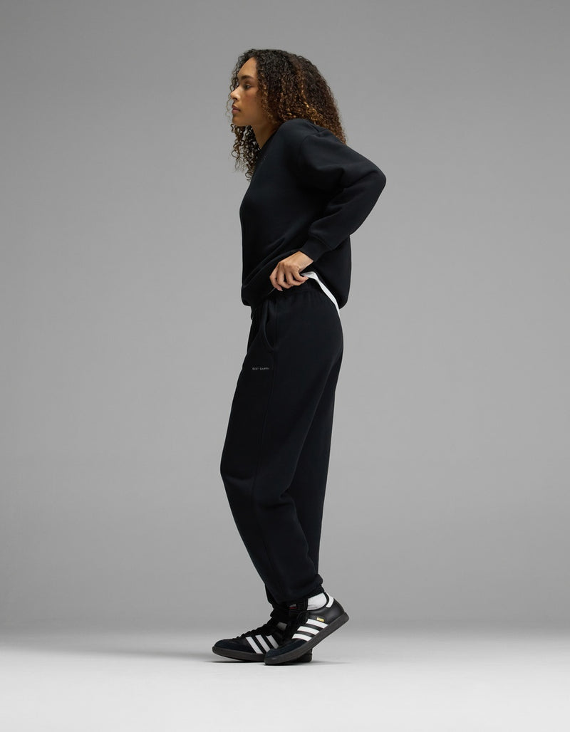 A person with curly hair stands sideways in a relaxed pose, wearing the Women’s CityScape Crewneck & Sweatpant Set by Cozy Earth in black, paired with black Adidas sneakers featuring white stripes, against a plain gray background.