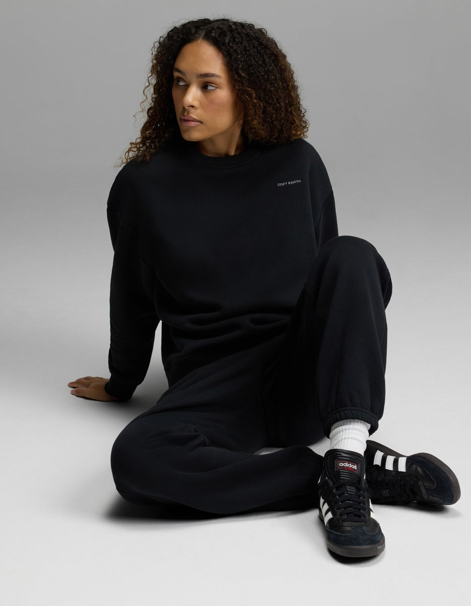 A woman with curly hair sits on the floor with one leg bent, looking to the side against a plain gray background, wearing the Women’s CityScape Crewneck & Sweatpant Set from Cozy Earth.