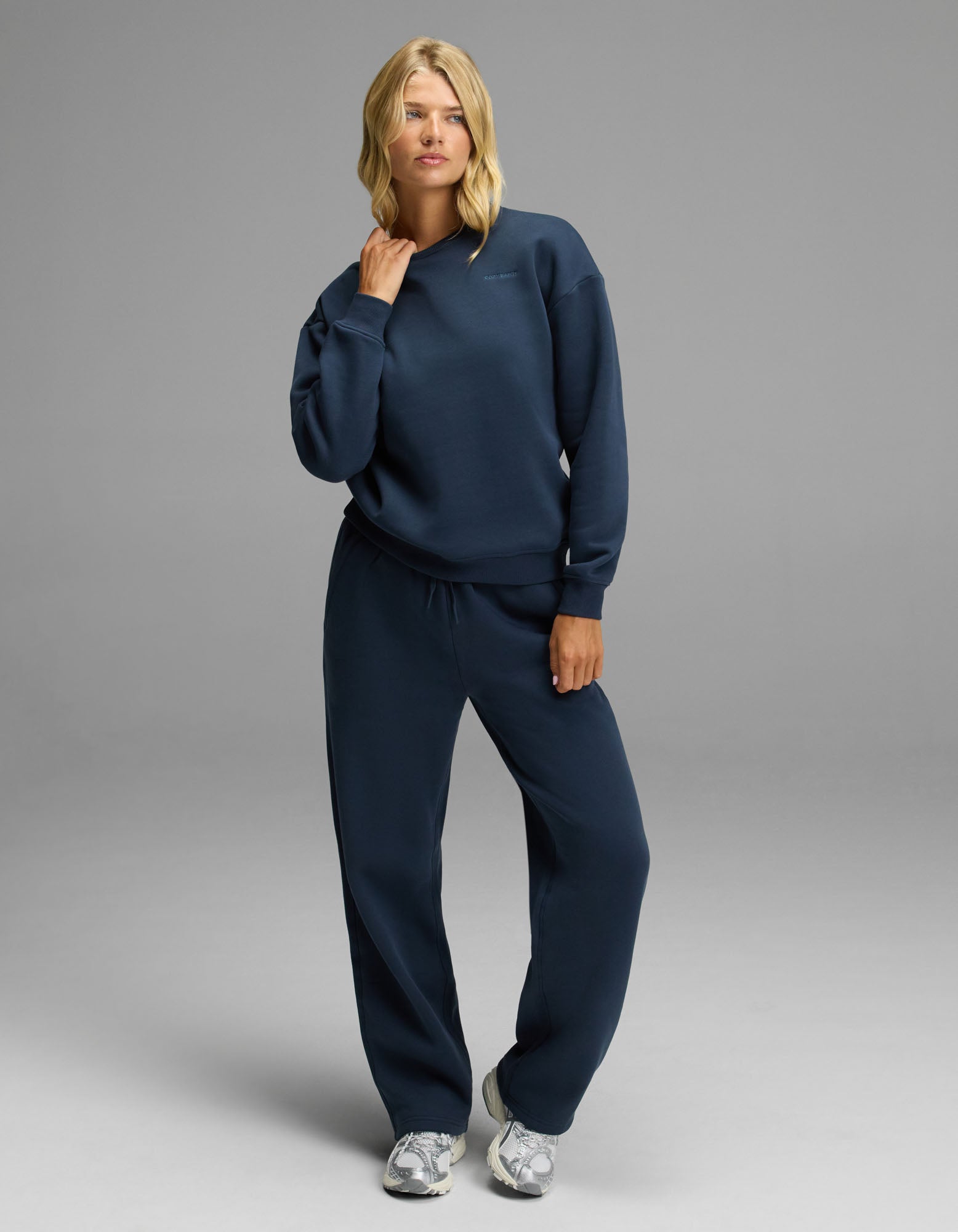 A woman with shoulder-length blonde hair stands against a gray background wearing the Cozy Earth Women’s Cityscape Crewneck & Wide Leg Pant Set in Eclipse, paired with silver sneakers. She holds her left hand near her chin.