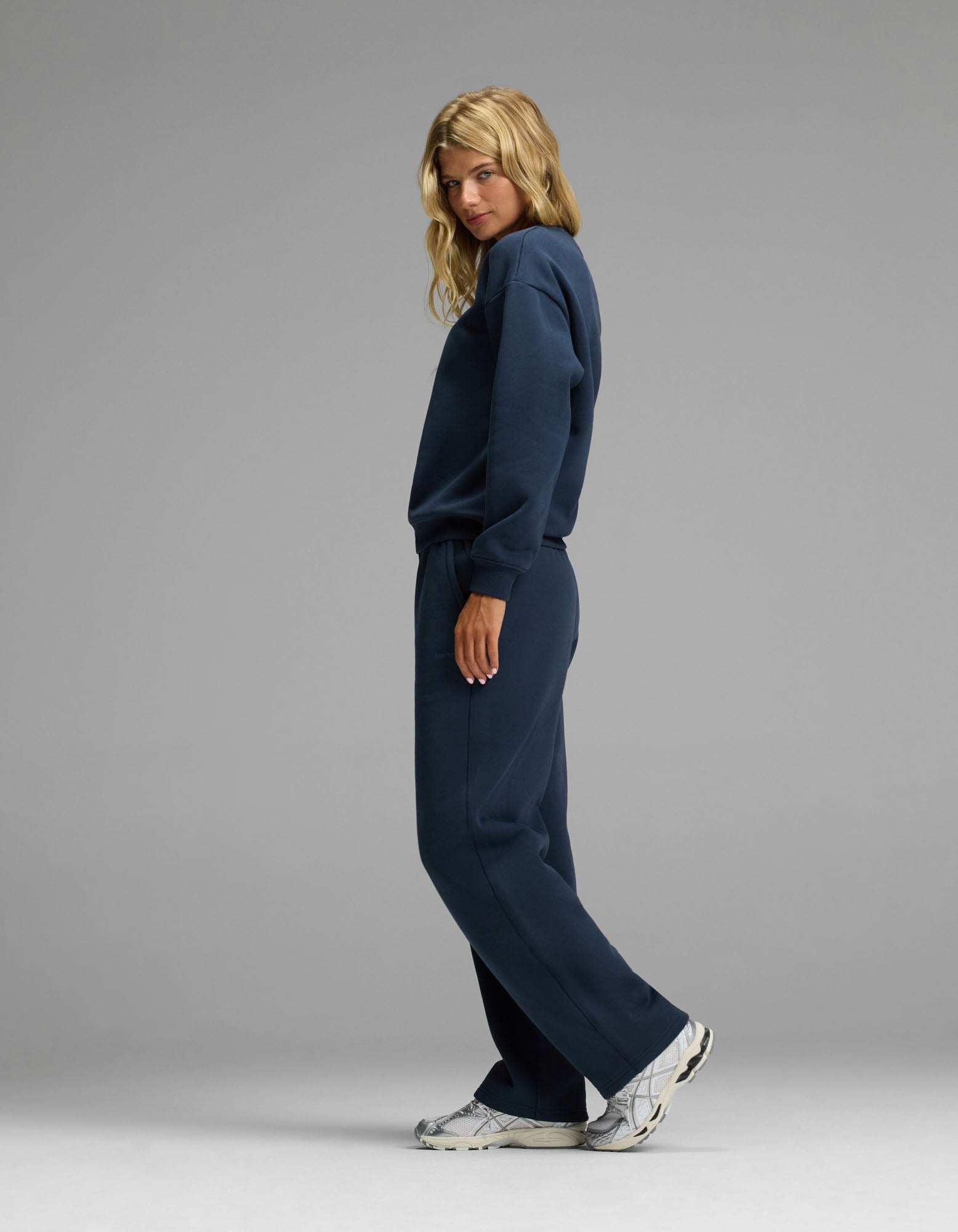 A person stands sideways, looking at the camera, wearing the Cozy Earth Women’s Cityscape Crewneck & Wide Leg Pant Set in navy blue with white sneakers, against a plain gray background.|Color:Eclipse