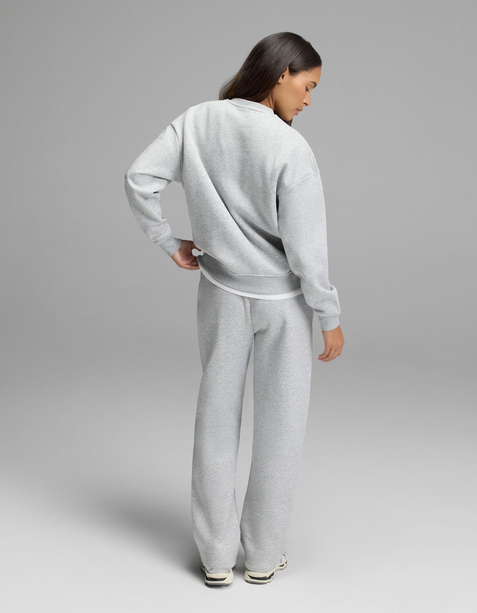 A person stands facing away, dressed in the Cozy Earth Women’s Cityscape 2.0 Crewneck & Wide Leg Pant Set in light gray with white sneakers, against a plain gray background.|Color:Heather Grey