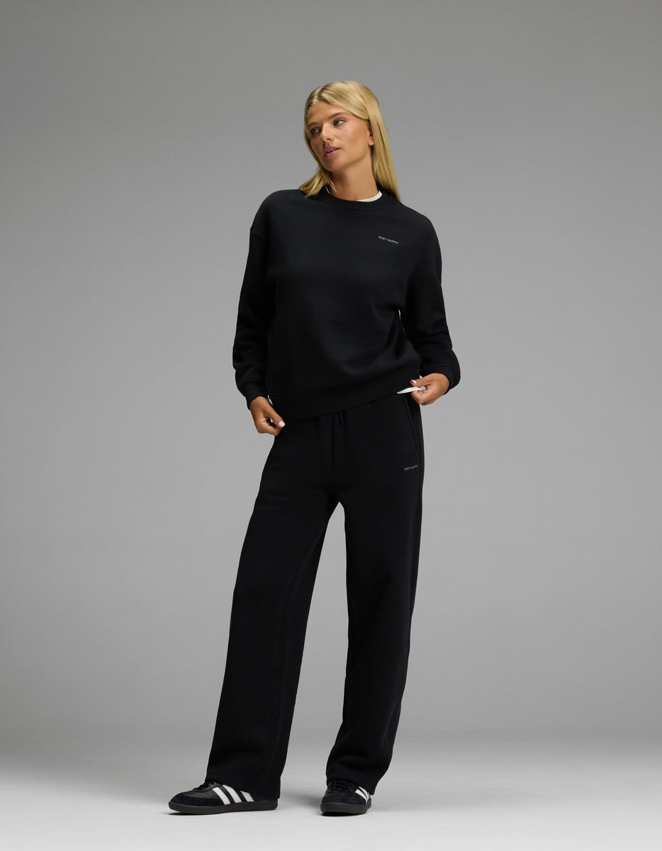 A person wearing the Cozy Earth Women’s CityScape Crewneck & Wide Leg Pant Set in black stands against a plain gray background, looking slightly to the side. They complete the look with black and white athletic shoes.