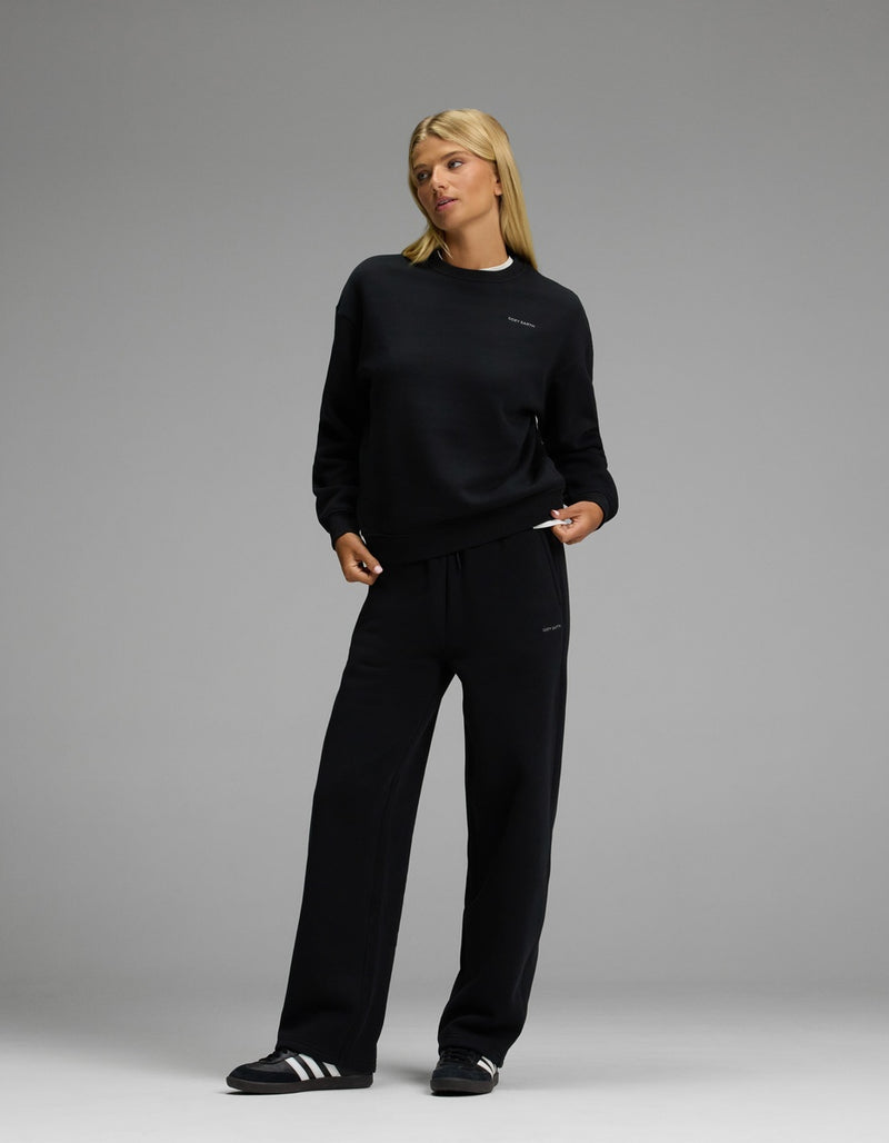 A person wearing the Cozy Earth Women’s CityScape Crewneck & Wide Leg Pant Set in black stands against a plain gray background, looking slightly to the side. They complete the look with black and white athletic shoes.
