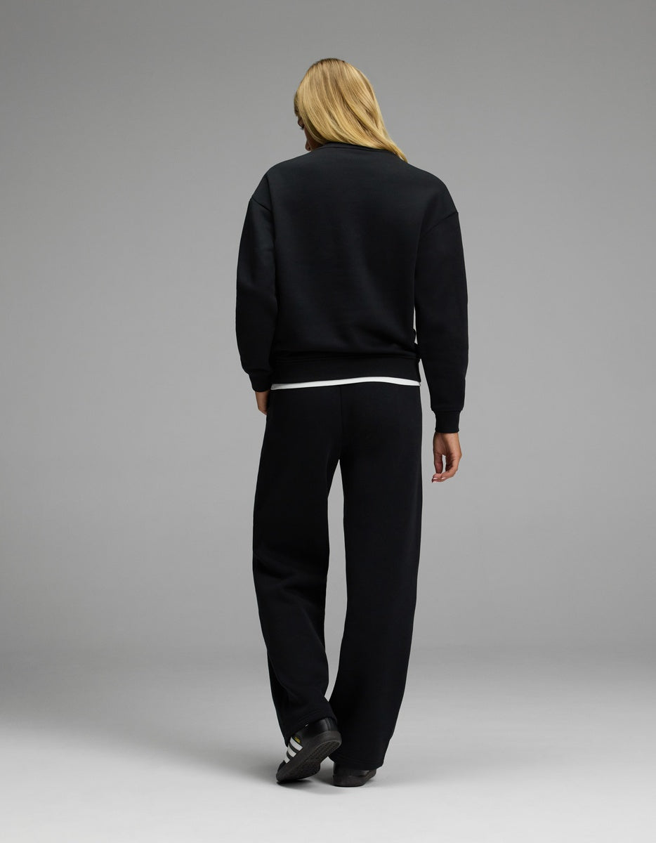 A person with long blonde hair stands facing away, wearing the Cozy Earth Women’s CityScape Crewneck & Wide Leg Pant Set in black, paired with black sneakers featuring white stripes, against a plain gray background.