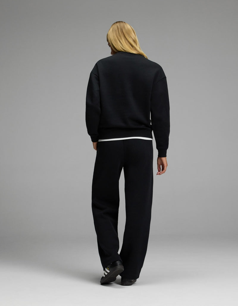 A person with long blonde hair stands facing away, wearing the Cozy Earth Women’s CityScape Crewneck & Wide Leg Pant Set in black, paired with black sneakers featuring white stripes, against a plain gray background.