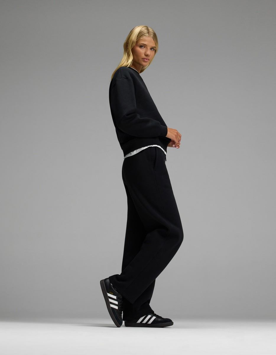 A person with long blonde hair wears Cozy Earth’s Women’s CityScape Crewneck & Wide Leg Pant Set in black, paired with black Adidas sneakers featuring white stripes, standing against a plain gray backdrop.