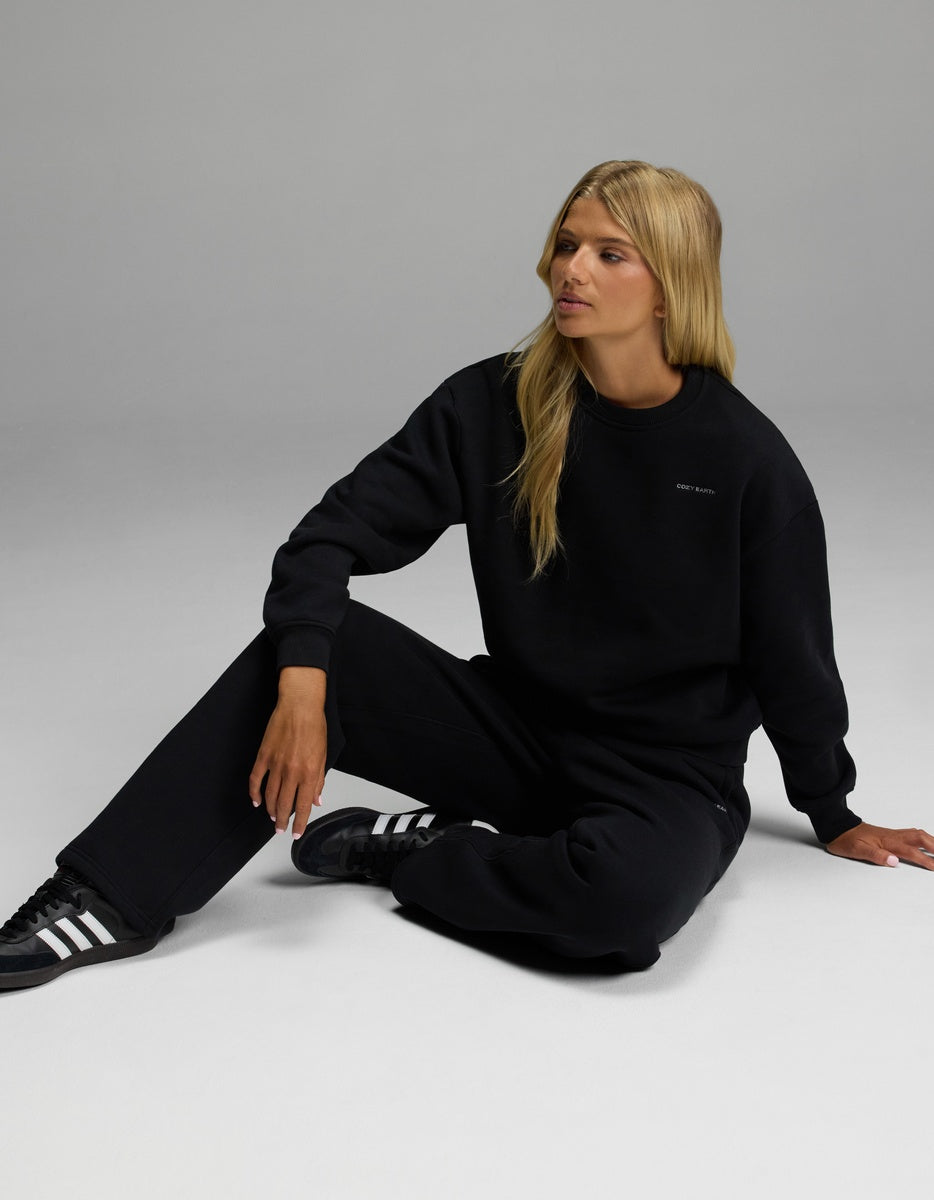 A person with long blonde hair sits on the floor wearing the Cozy Earth Women’s CityScape Crewneck & Wide Leg Pant Set in black, looking to the side against a plain light gray background.